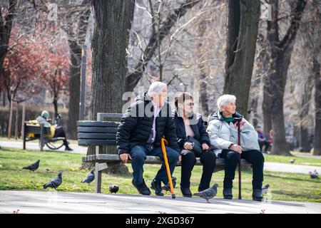 Plusieurs personnes retraitées sont assises sur un banc dans un parc public de la ville, profitant du temps ensoleillé, bavardant et se relaxant, entourées de verdure et d'arbres Banque D'Images