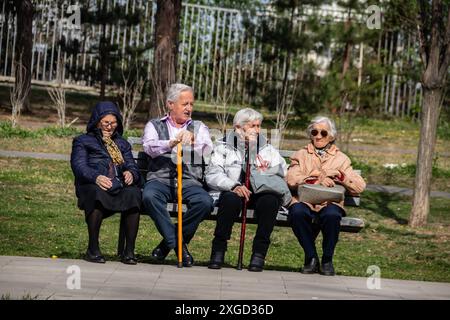 Plusieurs personnes retraitées sont assises sur un banc dans un parc public de la ville, profitant du temps ensoleillé, bavardant et se relaxant, entourées de verdure et d'arbres Banque D'Images