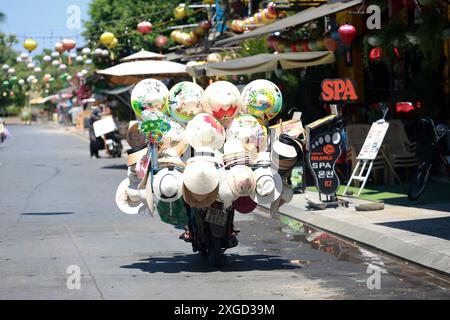 Hoi an, Vietnam 7 juillet 2024 : le colporteur vend ses chapeaux coniques vietness sur la moto. La vieille ville est un port commercial d'Asie du Sud-est datant du 15t Banque D'Images