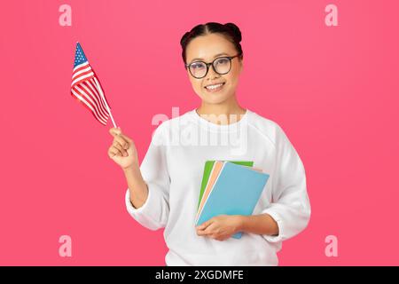 Femme souriante tenant drapeau américain et livres sur fond rose Banque D'Images