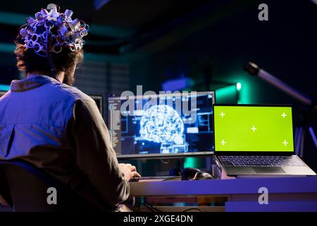 Ordinateur portable à écran isolé et homme avec casque EEG faisant le transfert du cerveau dans le monde virtuel, fusionnant avec l'intelligence artificielle. Admin utilisant les neurosciences et le Chroma Key Notebook pour gagner l'âme numérique Banque D'Images