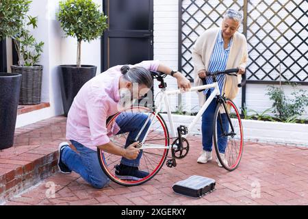 Fixation du vélo, homme senior agenouillé tandis que femme tenant le vélo dans la cour Banque D'Images