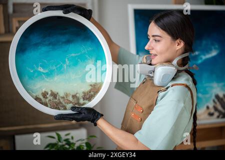Artiste féminine créative avec respirateur tient la peinture de résine époxy finie dans les mains appréciant le résultat. Banque D'Images