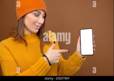 Portrait d'une jeune femme caucasienne attrayante dans un pull jaune et un chapeau tenant un smartphone. dans ses mains et pointe son doigt vers la découpe Banque D'Images