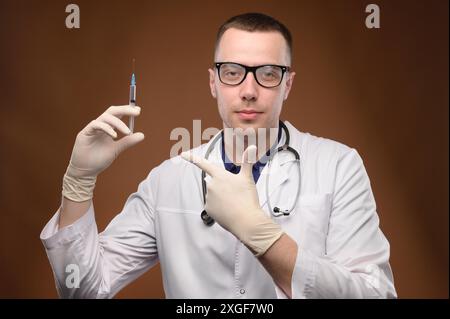 Un jeune médecin caucasien dans un manteau blanc et des lunettes tient une seringue avec un vaccin. Sympathique jeune homme câlin pointe un doigt sur une offre de seringue Banque D'Images