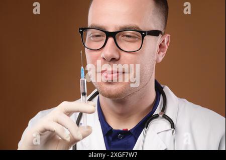 Bel homme en uniforme de médecin tenant une seringue souriant joyeusement, portrait studio shot Banque D'Images