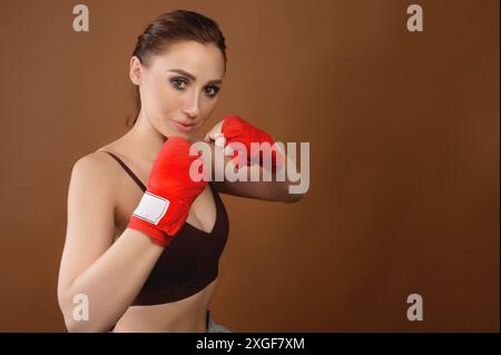 Une jeune femme en gants de boxe pose dans une position de combat et regarde la caméra. Boxeur féminin attrayant prêt à se battre sur fond brun Banque D'Images