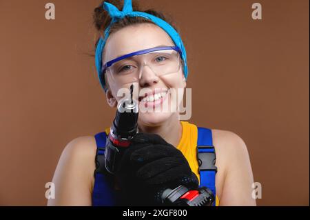 Mignonne fille dans des lunettes de construction protectrices presse un tournevis sur sa joue, constructeur heureux Banque D'Images