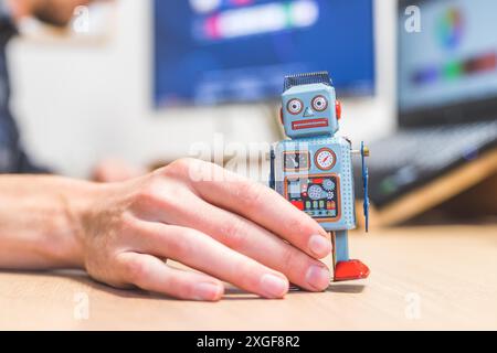 Robot jouet sur un bureau, homme travaillant sur l'ordinateur en arrière-plan. Symbole de l'intelligence artificielle, des chatbots ou des bots sociaux et des algorithmes Banque D'Images