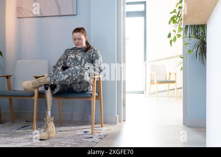 Assise dans la salle d'attente, femme en uniforme indéfini avec une jambe prothétique à l'aide d'un smartphone, espace de copie Banque D'Images