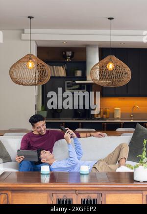 Détente à la maison, couple gay masculin riant et utilisant la tablette sur le canapé Banque D'Images