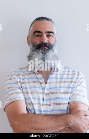 Homme senior barbu souriant avec les bras croisés portant une chemise rayée, à l'air confiant Banque D'Images