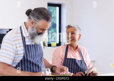 Couple aîné portant des tabliers cuisinant ensemble et souriant dans la cuisine Banque D'Images