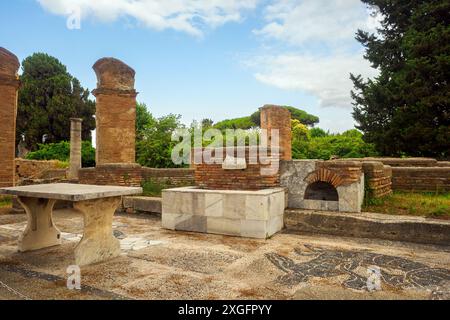 Macellum e Tabernae dei Pescivendoli - le Macellum est accessible depuis le Decumanus par une entrée avec des colonnes sur les côtés de laquelle, dans la première moitié du IIIe siècle après JC, deux tabernae (magasins) ont été créés, dotés de comptoirs pour la vente de poissons et de bassins en marbre. La salle de gauche conserve une mosaïque représentant un dauphin mangeant un calmar, avec l’inscription « Inbide te calco » (« créature envieuse, je te piétinerai »), car on pensait que les dauphins perturbaient la pêche.- Parc archéologique d’Ostie Antica, Rome, Italie Banque D'Images