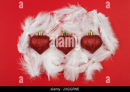3 bulbes de Noël en forme de coeur rouge sur des plumes blanches et un fond rouge Banque D'Images