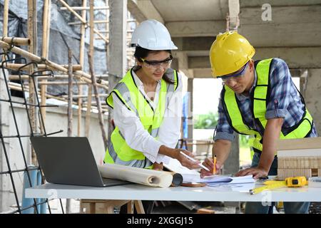 Architectes examinant les plans et les notes sur une table au chantier de construction avec ordinateur portable et modèle architectural Banque D'Images