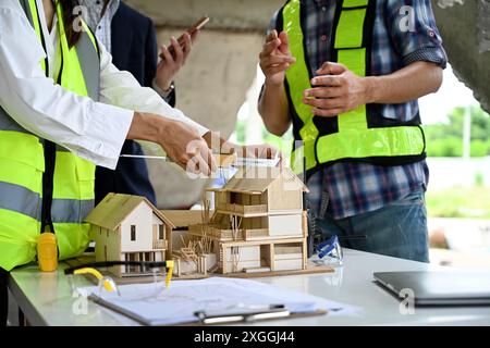 Architectes examinant les plans et les notes sur une table au chantier de construction avec ordinateur portable et modèle architectural Banque D'Images