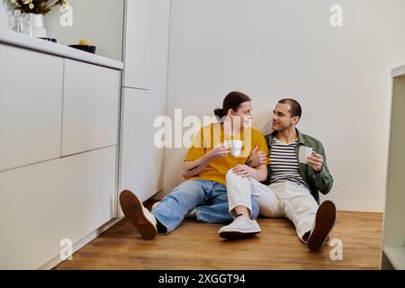 Un jeune couple gay est assis sur le sol de leur cuisine moderne, profitant d'une tasse de café et de l'autre compagnie. Banque D'Images