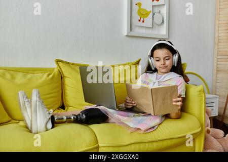 Une jeune fille avec une jambe prothétique se détend sur un canapé jaune, perdue dans un livre en portant des écouteurs. Banque D'Images