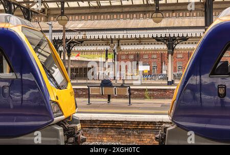 Deux locomotives se font face au quai d'une gare ferroviaire. Entre les deux et sur une autre plate-forme, un homme est assis sur un banc. Un bâtiment en briques est dans le di Banque D'Images