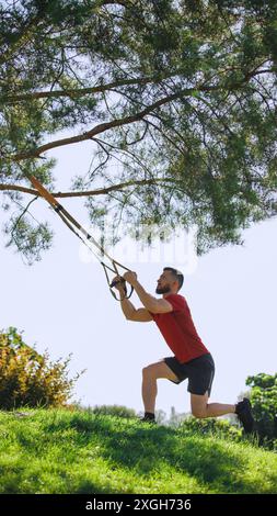 Homme athlétique faisant l'entraînement trx à l'extérieur, avec des sangles suspendues sur les branches d'arbre et faisant des squats. Séance d'entraînement active et intense Banque D'Images