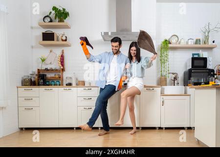 Jeune couple heureux ayant du plaisir tout en faisant le nettoyage de la cuisine, ils tiennent les chiffons avec flacon pulvérisateur dans les mains et heureusement à danser, sourire et rire toge Banque D'Images