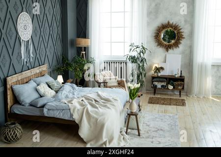 Chambre moderne avec lit en bois, draps bleus, décor en osier et plantes en pot. Grande fenêtre avec rideaux blancs fournit la lumière naturelle. Dreamcatcher Banque D'Images