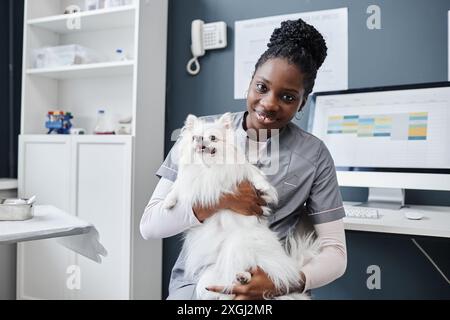 Portrait d'une jeune vétérinaire féminine d'origine afro-américaine portant des gommages gris tenant un chien blanc de poméranie mignon dans les mains regardant la caméra, dans le vétérinaire, le bureau, l'espace de copie Banque D'Images