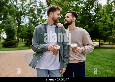 Deux hommes barbus, habillés de façon décontractée, se promènent main dans la main dans un parc verdoyant, profitant d’un moment partagé et d’une tasse de café. Banque D'Images