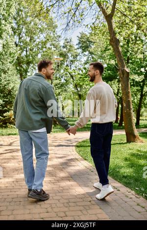 Un couple gay barbu marche main dans la main sur un chemin pavé dans un parc verdoyant, riant et profitant d'une journée ensoleillée ensemble. Banque D'Images