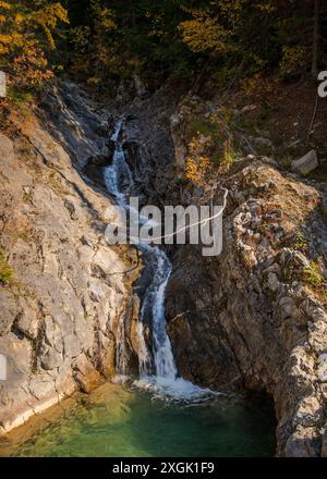 Petite cascade descendant un flanc de montagne rocheux en automne, créant un bassin d'eau verte au fond Banque D'Images