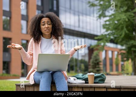 Jeune femme assise sur le banc sur le campus universitaire à l'aide d'un ordinateur portable avec une expression confuse. Tasse à café et sac à dos à côté d'elle. Frustration des élèves et concept d'apprentissage en plein air. Banque D'Images