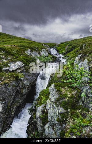 Un flanc de montagne rocheux en Norvège présente un ruisseau d'eau qui coule vers le bas, créant une cascade sous un ciel gris et nuageux Banque D'Images