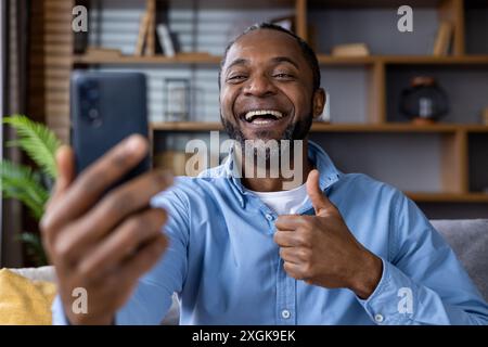 Homme heureux faisant un appel vidéo de la maison, souriant et donnant le pouce vers le haut geste. S'engager dans la communication virtuelle. Intérieur confortable de la maison en arrière-plan. Banque D'Images