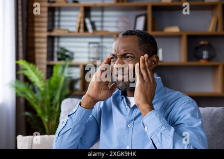 Homme stressé avec mal de tête assis sur le canapé dans le salon moderne. Il tient sa tête dans l'inconfort, indiquant la douleur et l'anxiété. Fond lumineux et bien organisé avec étagère et plantes. Banque D'Images