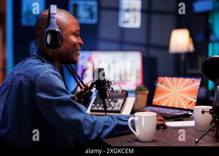Un homme noir souriant animant un talk-show est montré dans un profil de vue latérale, parlant avec ses auditeurs en ligne tout en faisant un enregistrement de podcast en direct depuis son home studio, il porte des écouteurs sans fil. Banque D'Images