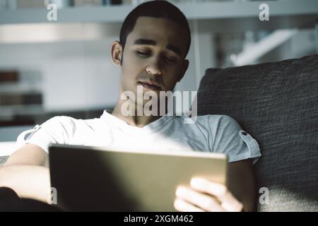 Jeune homme naviguant sur Internet sur sa tablette dans une maison moderne confortable, mettant en valeur l'intégration de la technologie dans la vie quotidienne Banque D'Images