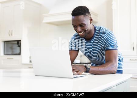 Heureux homme afro-américain utilisant un ordinateur portable s'appuyant sur le plan de travail dans la cuisine ensoleillée. Communication, rafraîchissement, matin, vie domestique et style de vie, inaltérable Banque D'Images