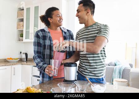 Couple gay biracial et moyen-oriental faisant des smoothies dans une cuisine à la maison. Ils partagent un moment joyeux tout en préparant une boisson saine ensemble. Banque D'Images