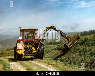 Un tracteur équipé d'un accessoire à lame rotative tond un flanc de coteau, laissant un andain d'herbe coupée dans son sillage. Le tracteur se déplace sur un chemin de terre avec une vue o Banque D'Images