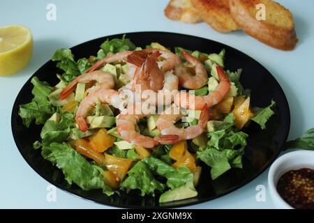 Une salade de crevettes fraîches avec avocat et légumes verts sur une assiette noire, accompagnée de citron, pain grillé, et un petit bol de vinaigrette Banque D'Images