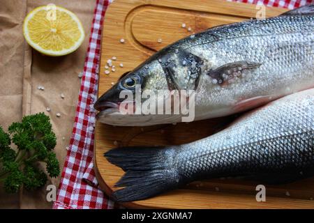 Un poisson cru frais repose sur une planche à découper en bois à côté d'un citron à moitié coupé, persil et sel, le tout sur un papier brun et un tissu à carreaux rouge Banque D'Images