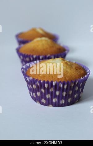 Trois cupcakes jaunes dans des doublures en papier à pois violets disposés en rangée sur un fond blanc Banque D'Images