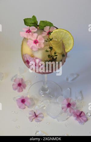 Un cocktail rafraîchissant garni d'une tranche de citron, de fleurs roses et de feuilles de menthe, entouré de glace et de fleurs supplémentaires Banque D'Images