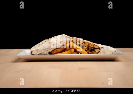 Un wrap grillé rempli de poulet et de légumes servis avec des rondelles d'oignon sur une assiette blanche sur une table en bois sur fond noir Banque D'Images