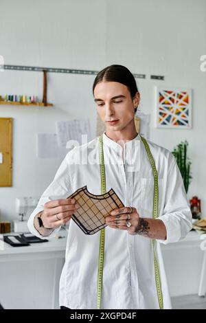 Un jeune homme en chemise blanche, avec un ruban à mesurer autour du cou, examine un morceau de tissu dans son atelier de restauration de vêtements. Banque D'Images