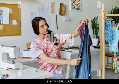 Un jeune homme, vêtu d’une chemise teinte au noeud, examine un morceau de tissu denim dans son atelier de restauration de vêtements. Banque D'Images