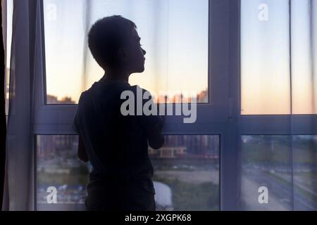 Silhouette de garçon debout à la fenêtre à l'aube regardant quelque chose d'intéressant. Vie urbaine. Banque D'Images