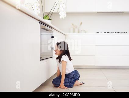 Cuisson, four ou attente et fille dans la cuisine de la maison pour la patience à la cuisson des aliments avec l'appareil. Excité, heureux et sourire de l'enfant boulanger sur le sol de Banque D'Images