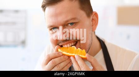 Portrait de Chef professionnel Bite Orange Slice. Homme Vlogger mangeant jus Citrus avec sourire sur le visage. Personne a mis des fruits jaunes à la bouche. Des vies alimentaires saines Banque D'Images